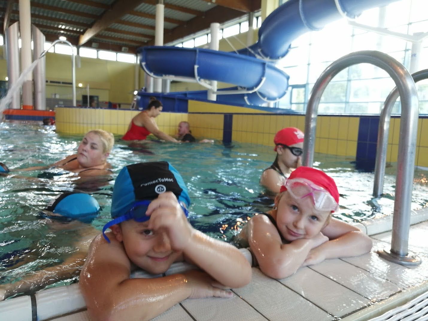 Dwoje uśmiechniętych dzieci odpoczywa przy krawędzi basenu po zajęciach nauki pływania, w tle zjeżdżalnia wodna i inni uczestnicy.