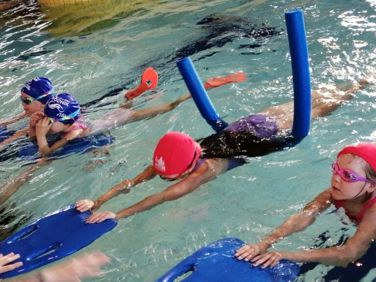 Dzieci uczące się pływać w basenie, korzystające z makaronów i desek do nauki pływania, ubrane w kolorowe czepki i okularki.