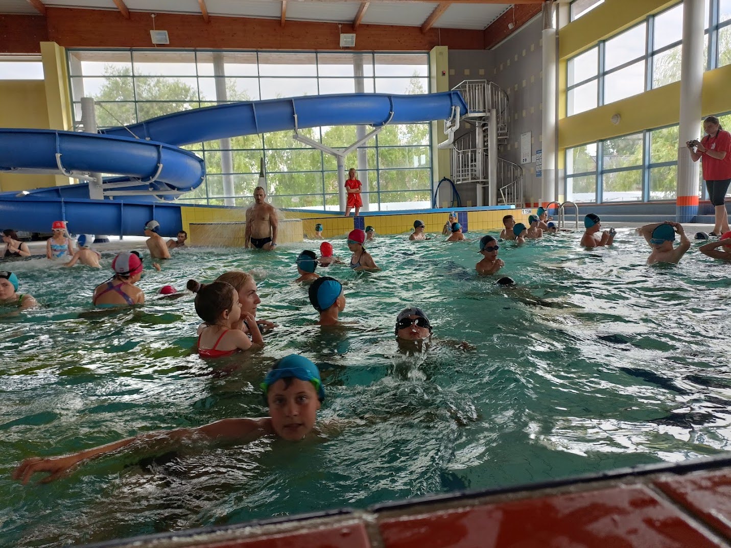 Liczne dzieci podczas zajęć na basenie, pływające i bawiące się pod opieką instruktorów w dużym, krytym aquaparku z niebieską zjeżdżalnią.