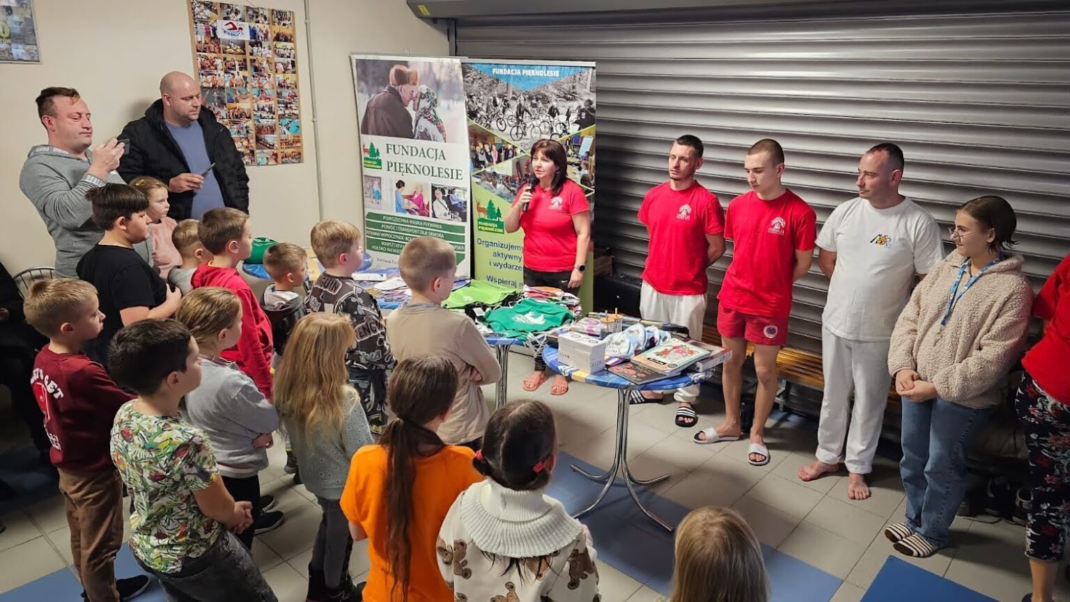 Grupa dzieci i dorosłych zgromadzona wokół stolika z nagrodami i materiałami promocyjnymi Fundacji Pięknolesie, podczas podsumowania projektu na basenie.
