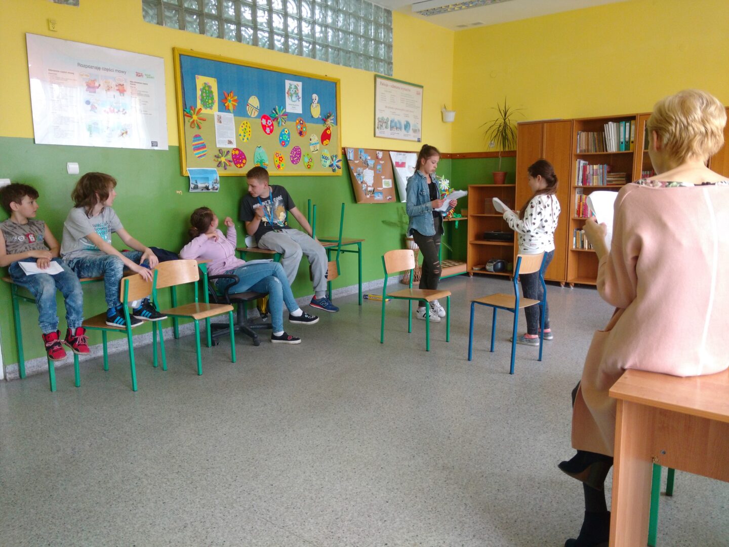 Grupa dzieci uczestnicząca w warsztatach teatralnych – część siedzi, obserwując, a część występuje na środku sali, trzymając scenariusze w rękach.