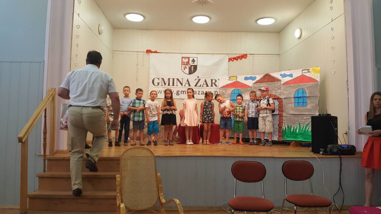 Grupa dzieci uczestnicząca w warsztatach teatralnych – część siedzi, obserwując, a część występuje na środku sali, trzymając scenariusze w rękach.