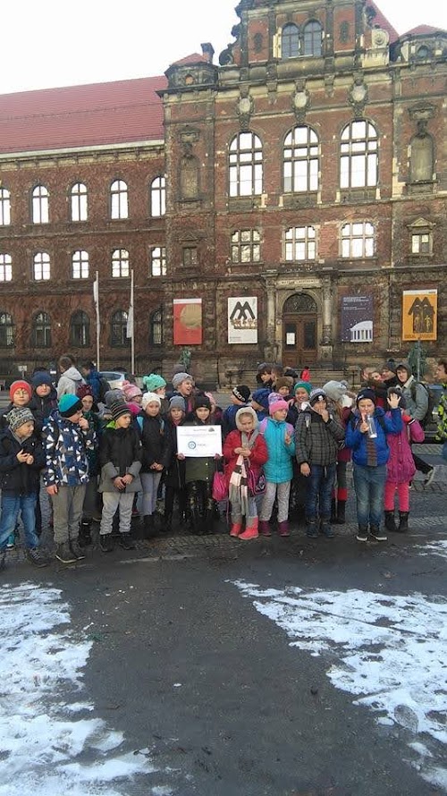 Grupa dzieci ubrana w zimowe kurtki i czapki pozuje na tle zabytkowego budynku muzeum podczas edukacyjnej wycieczki w ramach projektu kulturalnego.