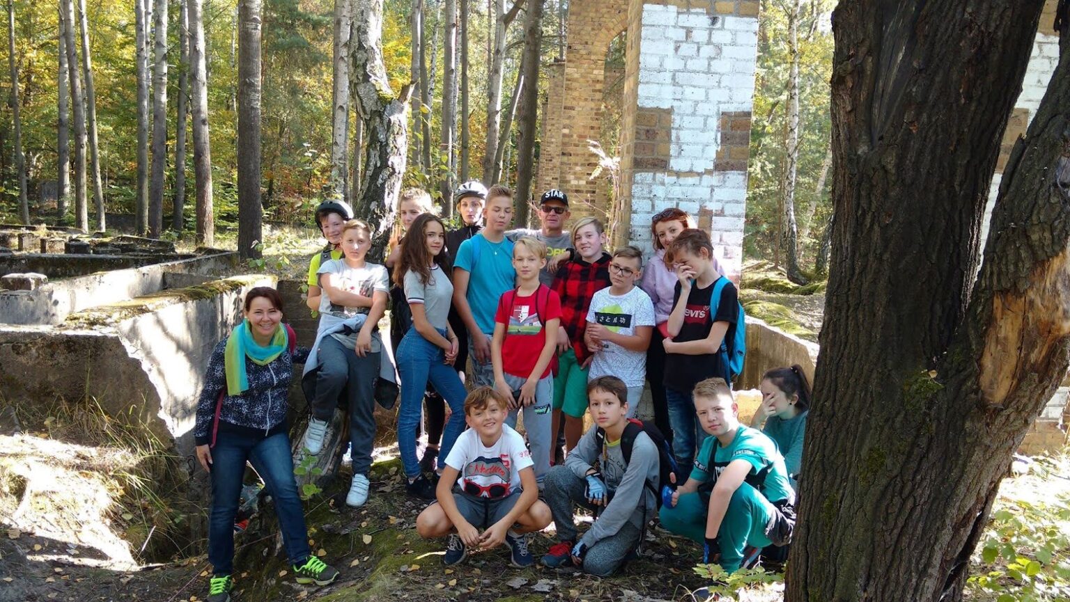 Grupa młodzieży podczas wizyty studyjnej w lesie przy ruinach, otoczona drzewami i śladami dawnej zabudowy, pozuje do zdjęcia w trakcie projektu edukacyjno-turystycznego.