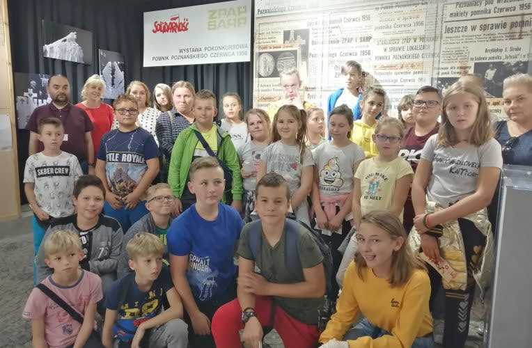 Grupa dzieci i młodzieży podczas wizyty w muzeum w Poznaniu, stojąca na tle ekspozycji dotyczącej wydarzeń Czerwca 1956 roku.