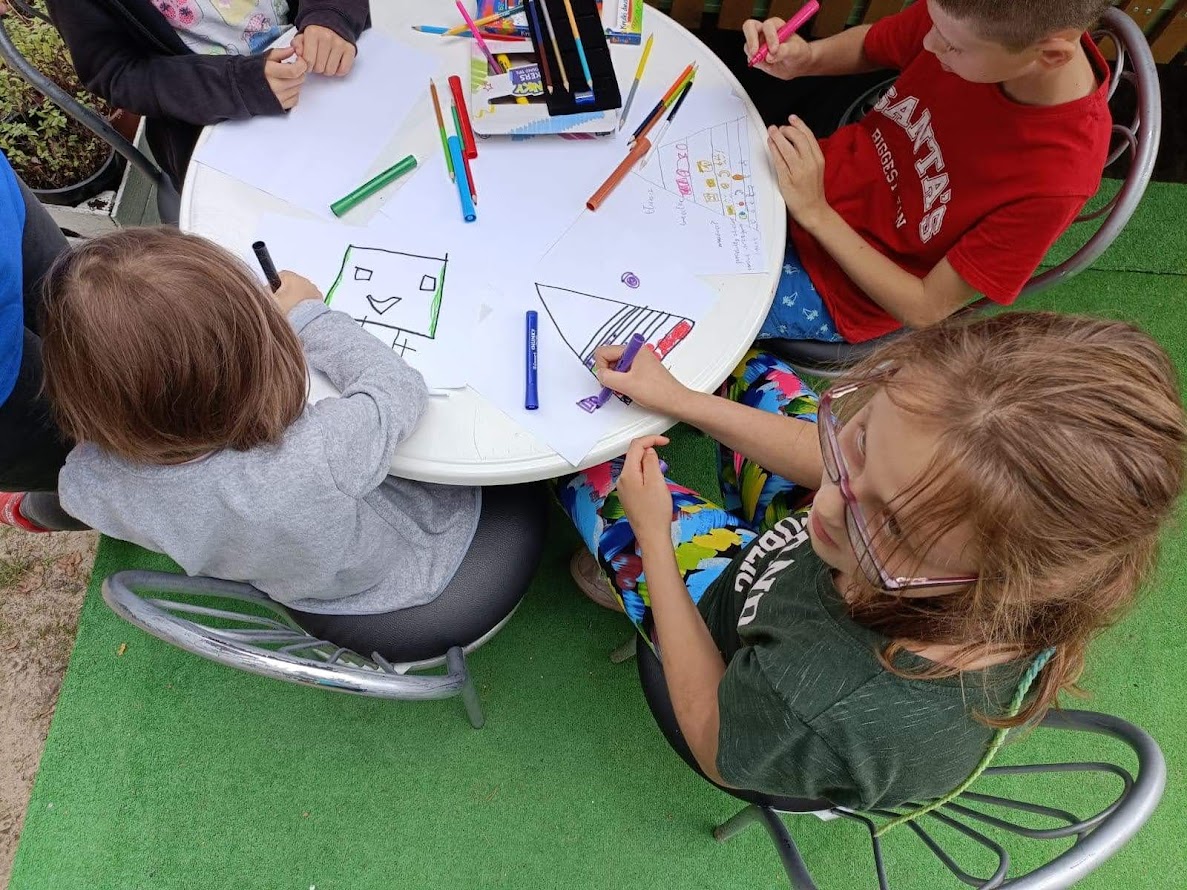 Dzieci rysujące kredkami i pisakami przy okrągłym stole podczas plenerowych warsztatów artystycznych.