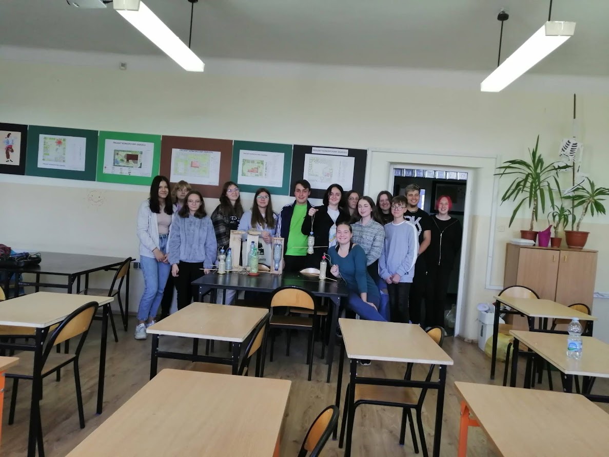 Grupa młodzieży pozująca wspólnie w sali lekcyjnej podczas warsztatów ekologicznych, za nimi plansze z projektami zielonych przestrzeni.