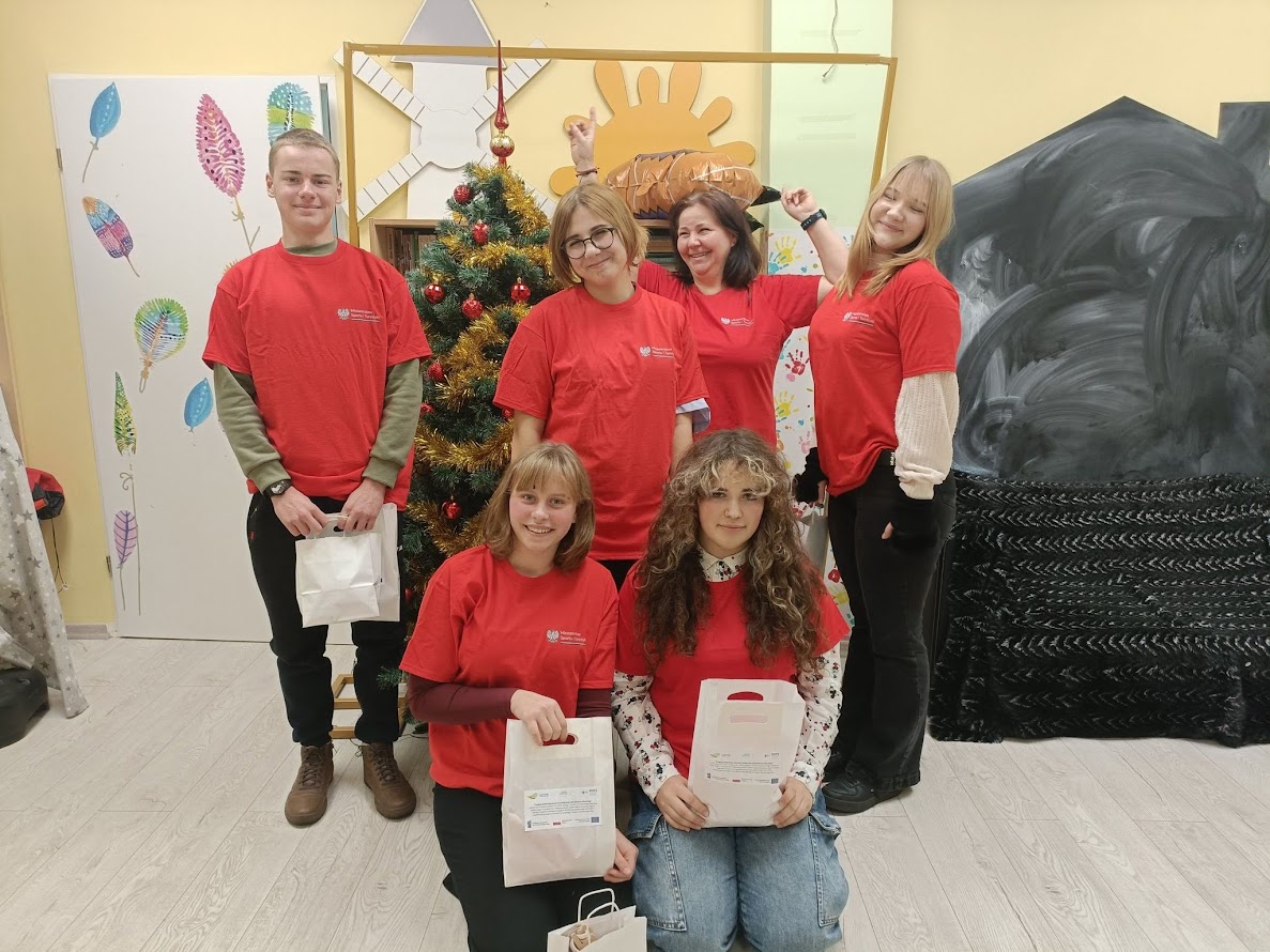 Grupa młodych wolontariuszy w czerwonych koszulkach pozuje do zdjęcia przy choince w udekorowanej sali, trzymając upominki.