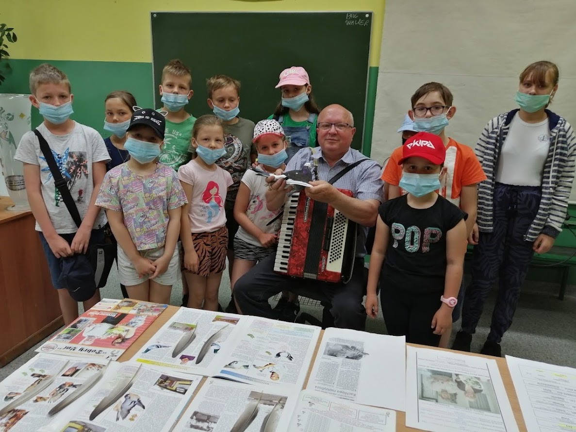 Dzieci w maseczkach ochronnych pozują z prowadzącym zajęcia mężczyzną grającym na akordeonie. Przed nimi leżą materiały edukacyjne związane z ptakami.