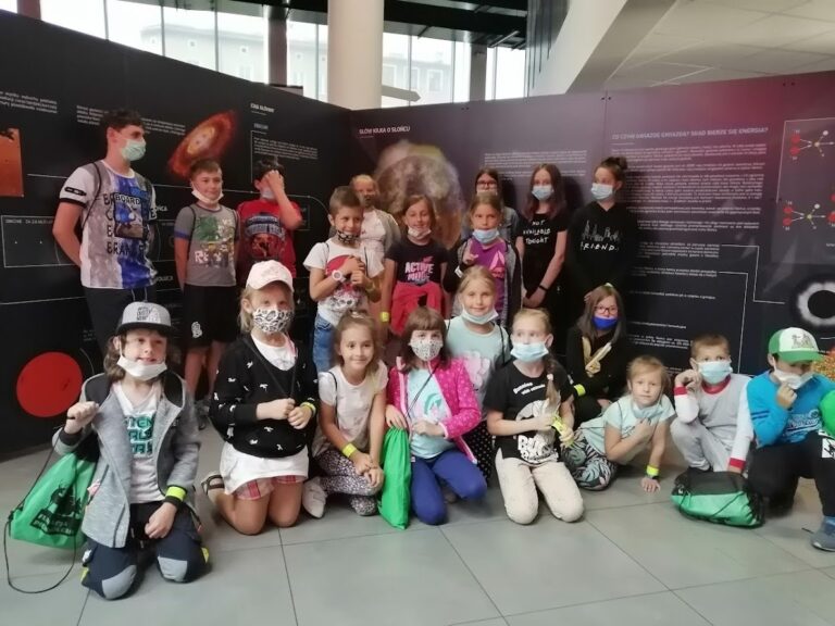 Grupa dzieci i młodzieży w maseczkach ochronnych pozuje do zdjęcia na tle tablic edukacyjnych o kosmosie, przedstawiających informacje o gwiazdach i słońcu.