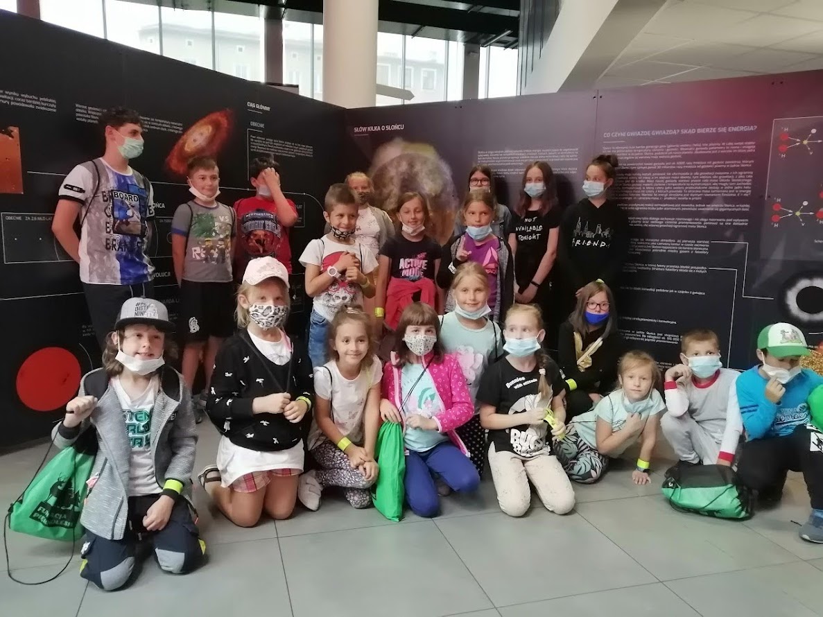 Grupa dzieci i młodzieży w maseczkach ochronnych pozuje do zdjęcia na tle tablic edukacyjnych o kosmosie, przedstawiających informacje o gwiazdach i słońcu.
