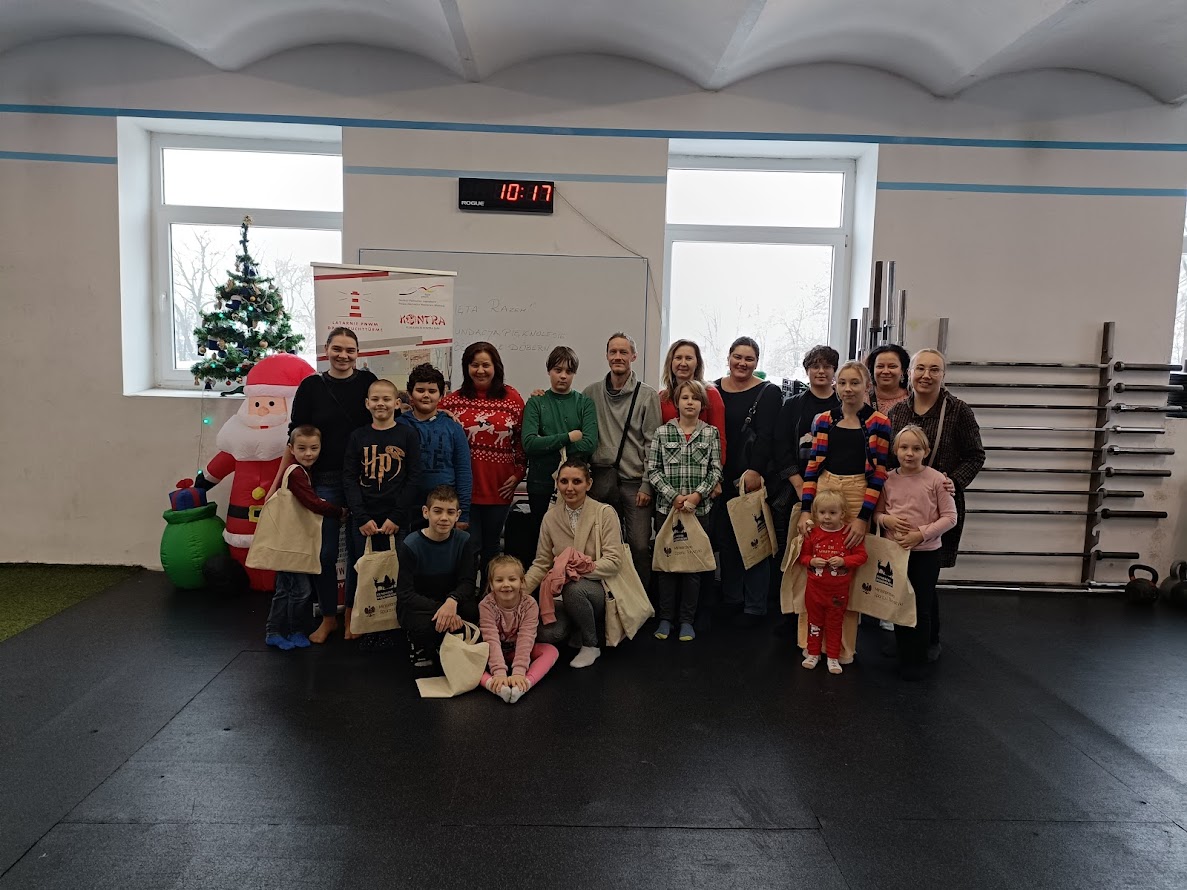 Grupa dzieci i dorosłych pozuje wspólnie do świątecznego zdjęcia na sali sportowej, obok udekorowanej choinki i dmuchanej postaci Mikołaja. Wszyscy trzymają bawełniane torby z prezentami.