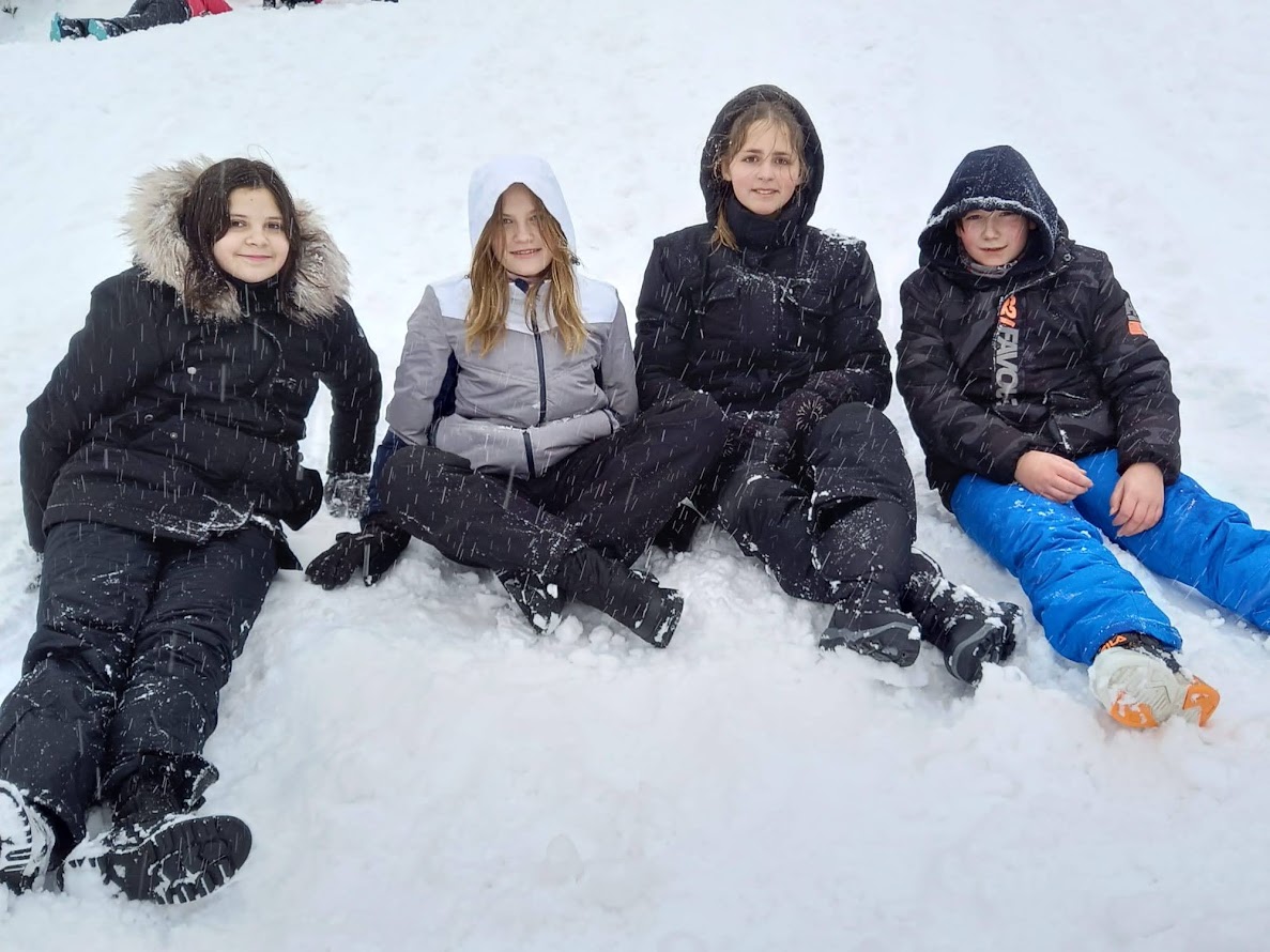 Czwórka dzieci ubrana w zimowe kurtki i spodnie siedzi na śniegu podczas zimowego wyjazdu. W tle widać padający śnieg i białe stoki.