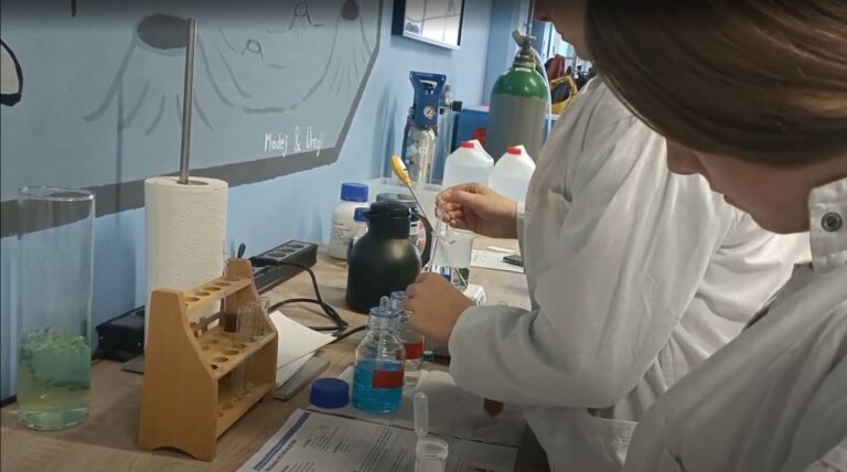 Dwie osoby w białych fartuchach laboratoryjnych przeprowadzają eksperymenty chemiczne przy stole wyposażonym w probówki, pipety i odczynniki.