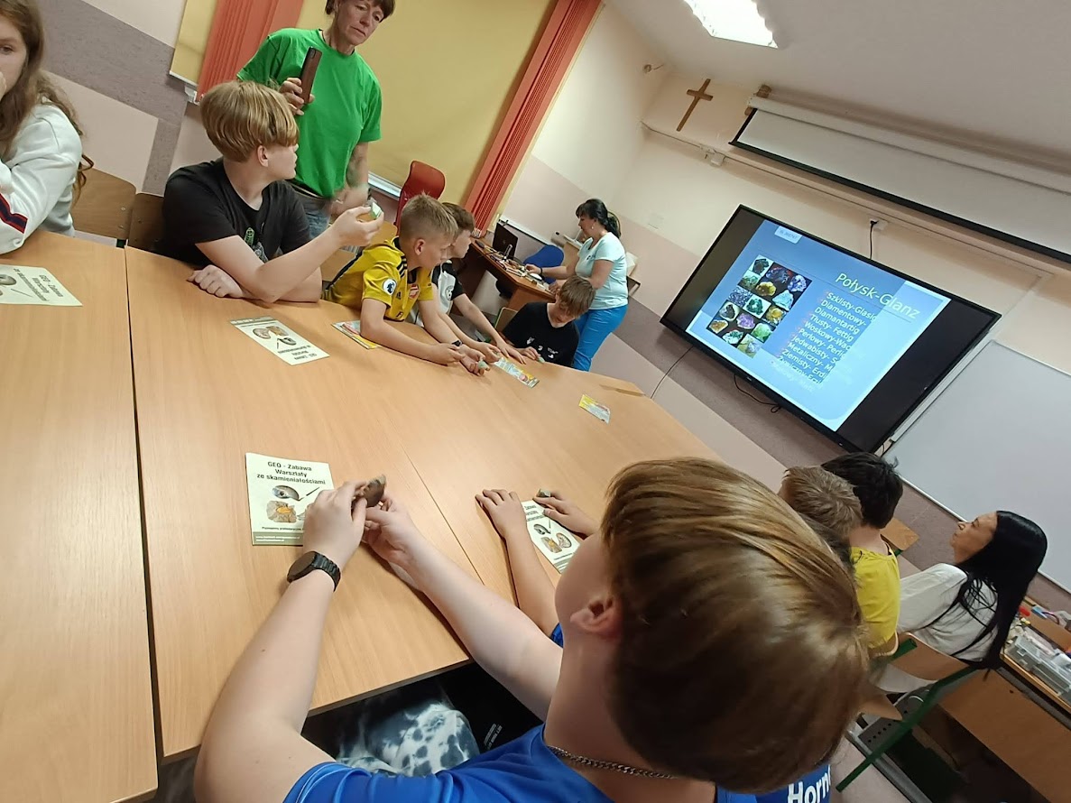 Grupa młodzieży siedzi przy stołach w klasie, uczestnicząc w warsztatach o minerałach. Na ekranie wyświetlony jest slajd z kolorowymi minerałami i napisami w języku polskim i niemieckim.