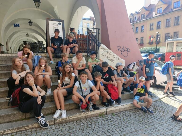 Grupa młodzieży odpoczywa na schodach pod arkadami w centrum miasta, jedząc lody i pijąc napoje podczas przerwy w programie wycieczki.