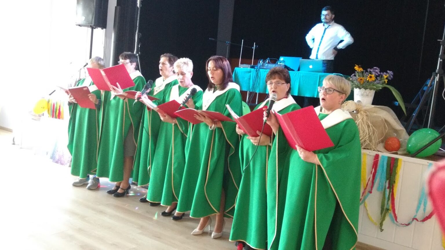 Grupa seniorek ubrana w zielone chóralne stroje występuje na scenie, śpiewając z czerwonych śpiewników podczas wydarzenia muzycznego.