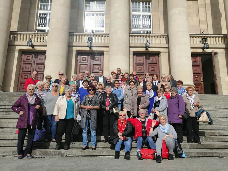 Grupa seniorek i seniorów pozuje do wspólnego zdjęcia na schodach reprezentacyjnego budynku w trakcie wyjazdu integracyjnego w ramach projektu.