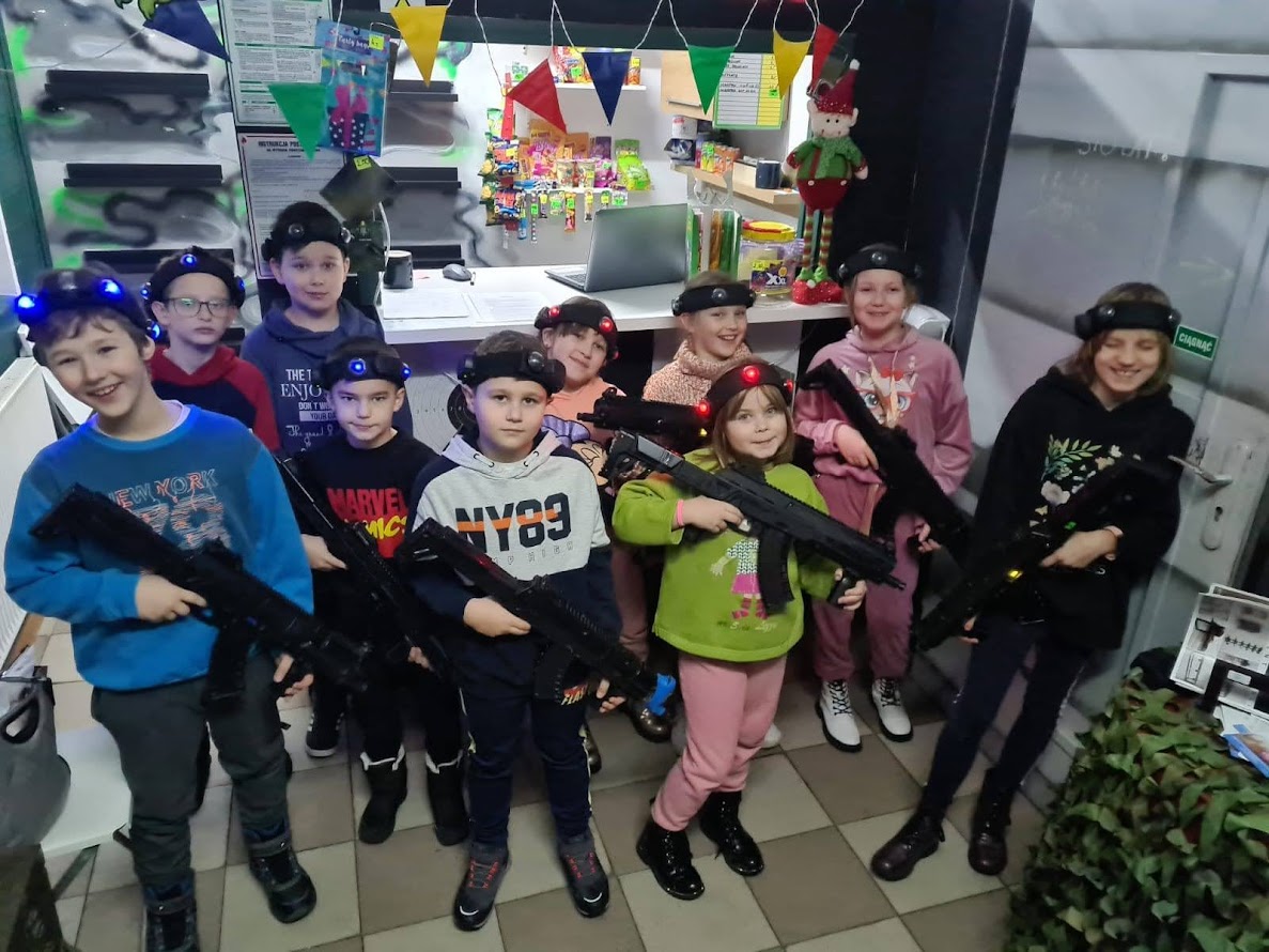 Grupa dzieci w kolorowych ubraniach trzymających sprzęt do gry w laserowy paintball, pozujących uśmiechniętych w centrum rozrywki.