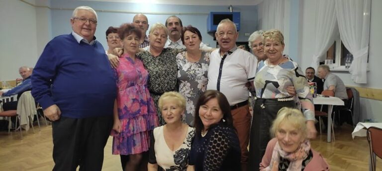 Grupa uśmiechniętych seniorów i seniorek pozuje wspólnie do zdjęcia podczas spotkania integracyjnego w sali z jasnymi ścianami i zasłonami.