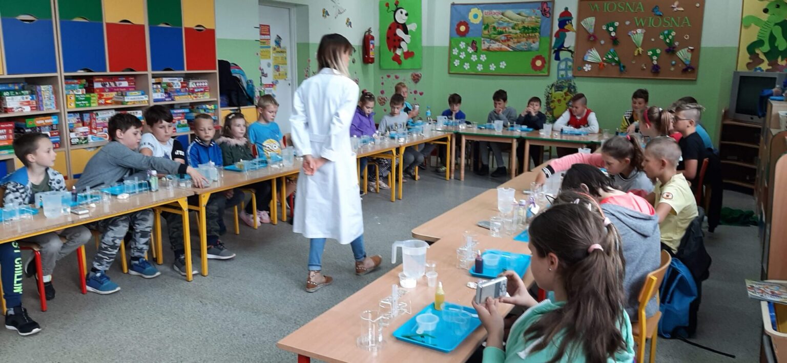 Grupa dzieci uczestniczy w warsztatach eksperymentalnych z prowadzącą ubraną w biały fartuch, w sali szkolnej udekorowanej planszami edukacyjnymi.
