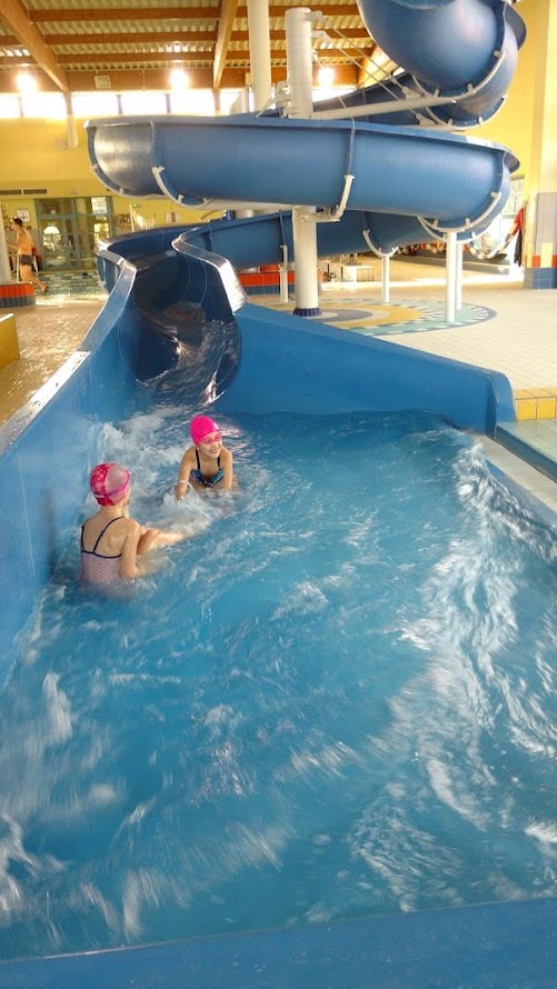 Dwie dziewczynki w różowych czepkach zjeżdżają z wodnej zjeżdżalni na basenie podczas zajęć pływackich.