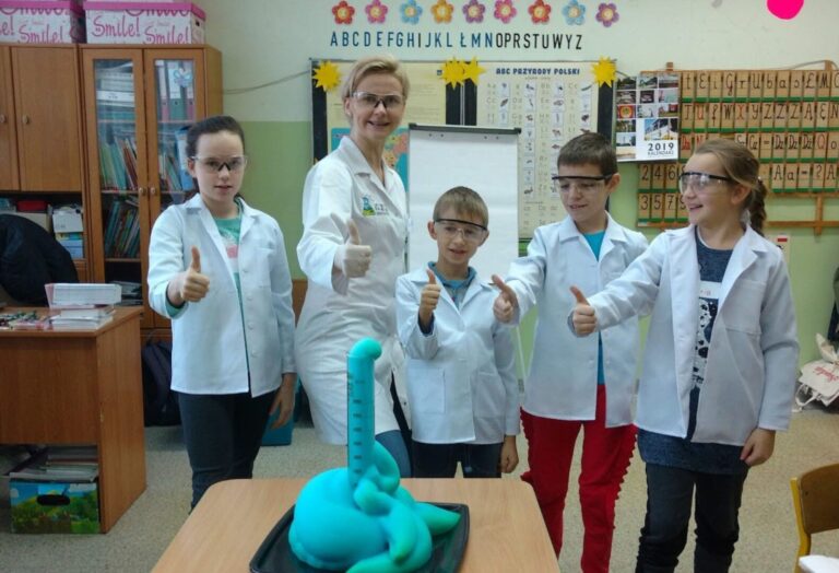 Grupa dzieci w białych fartuchach i okularach ochronnych pozuje z instruktorką przy kolorowym eksperymencie chemicznym w klasie.