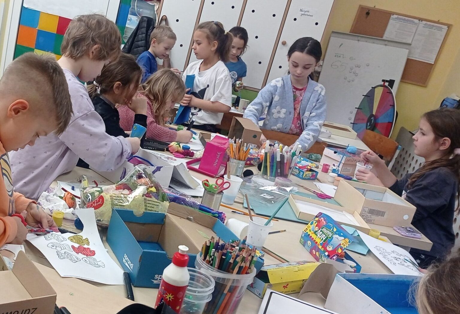 Dzieci i młodzież pracują przy stole, wykonując świąteczne ozdoby i kartki w ramach wspólnego projektu.