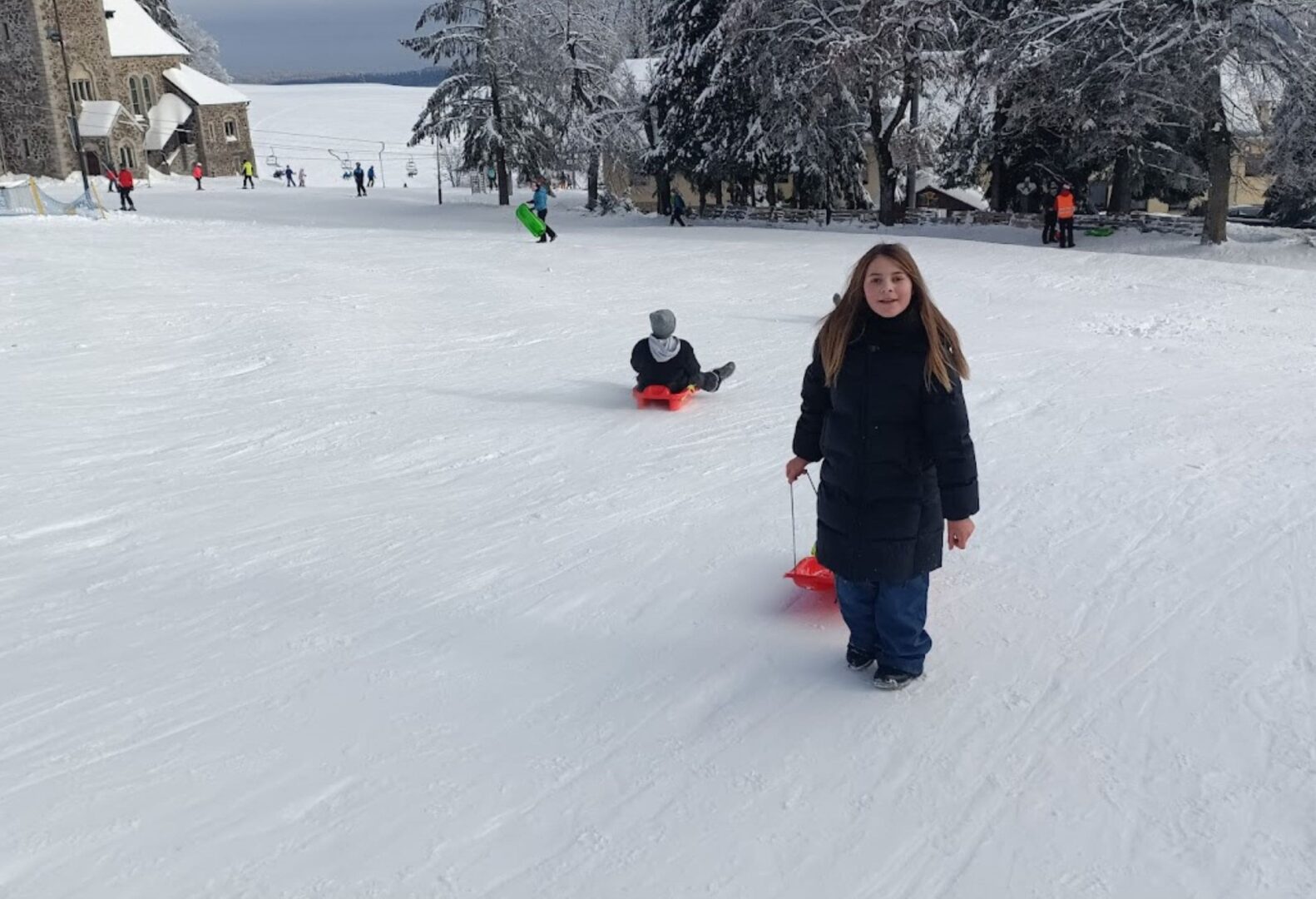 Dziewczynka w czarnej kurtce ciągnie plastikowy ślizgacz po ośnieżonym stoku; w tle dzieci zjeżdżają na sankach.