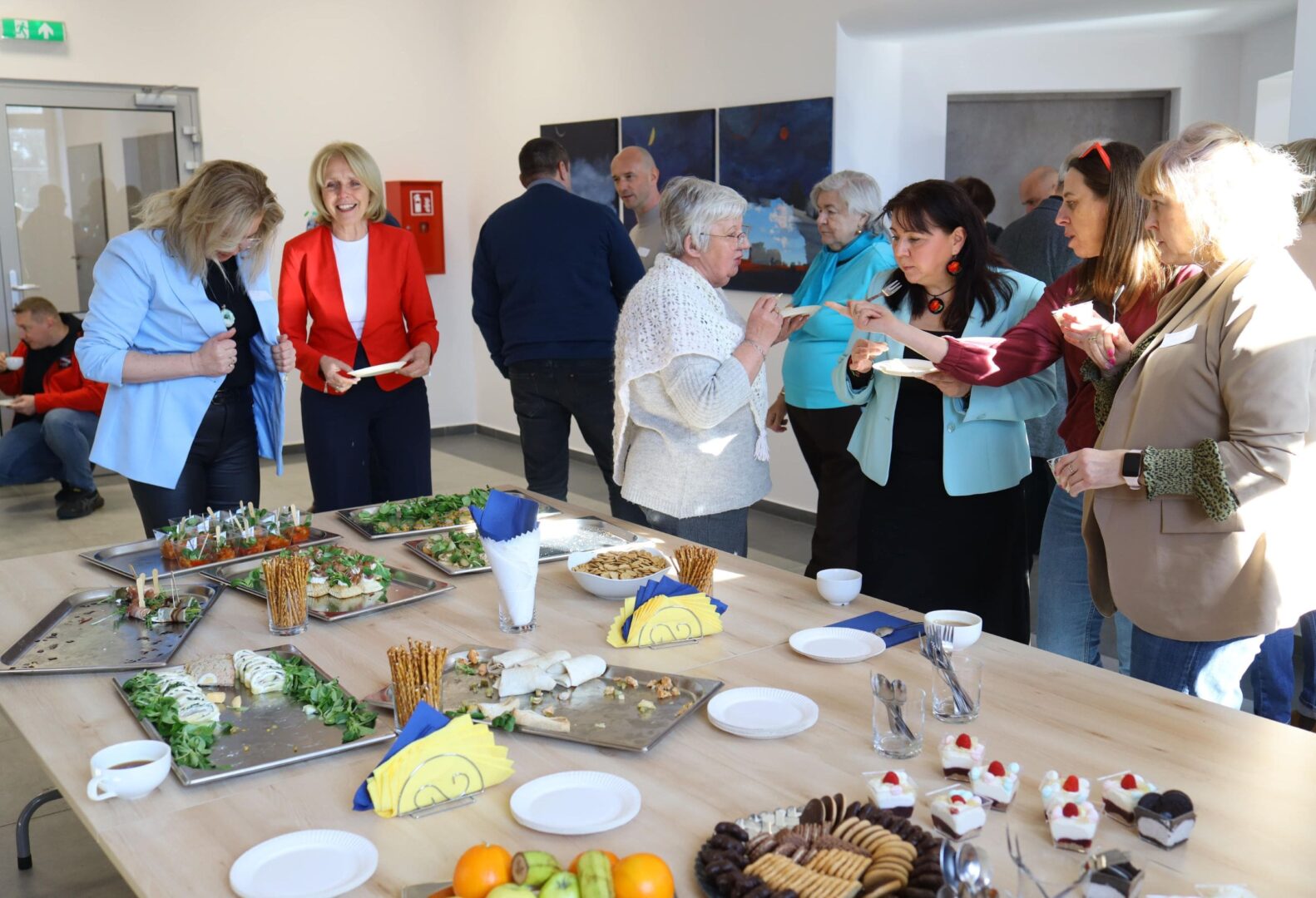 Uczestnicy imprezy integracyjnej Fundacji Pięknolesie rozmawiają przy stole zastawionym przekąskami i deserami.