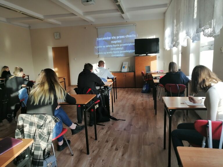 Uczniowie siedzą w klasie przy biurkach, notując treści z prezentacji wyświetlanej na ekranie.