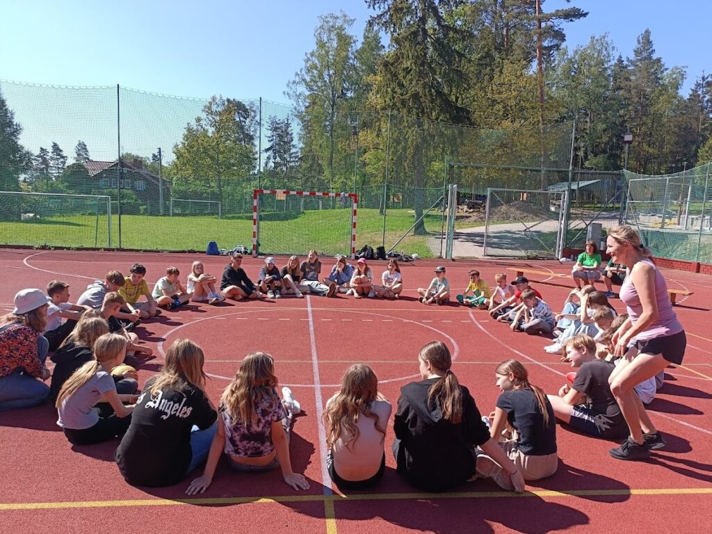 Grupa dzieci i młodzieży siedzi w kręgu na boisku, uczestnicząc w integracyjnych zajęciach na świeżym powietrzu.