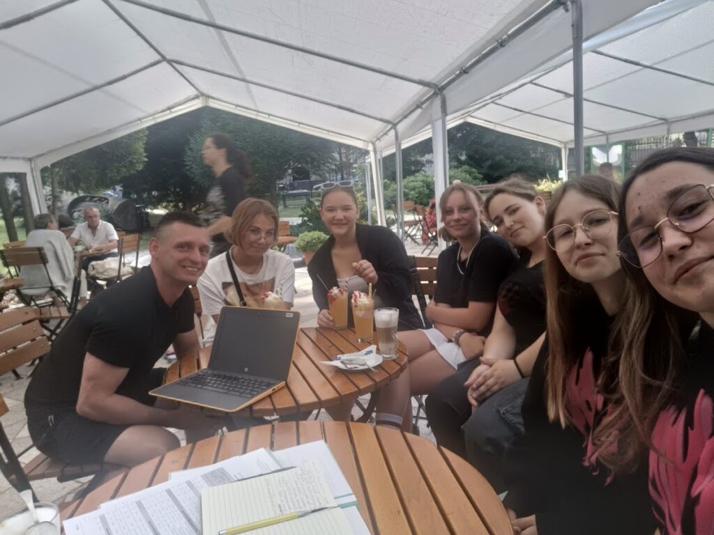 Grupa młodzieży i prowadzący siedzą przy stoliku w plenerowej kawiarni, rozmawiając i pracując z laptopem oraz notatkami.