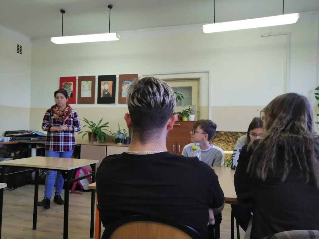 Grupa młodzieży siedzi w klasie i słucha prowadzącej warsztaty kobiety stojącej przy biurku.