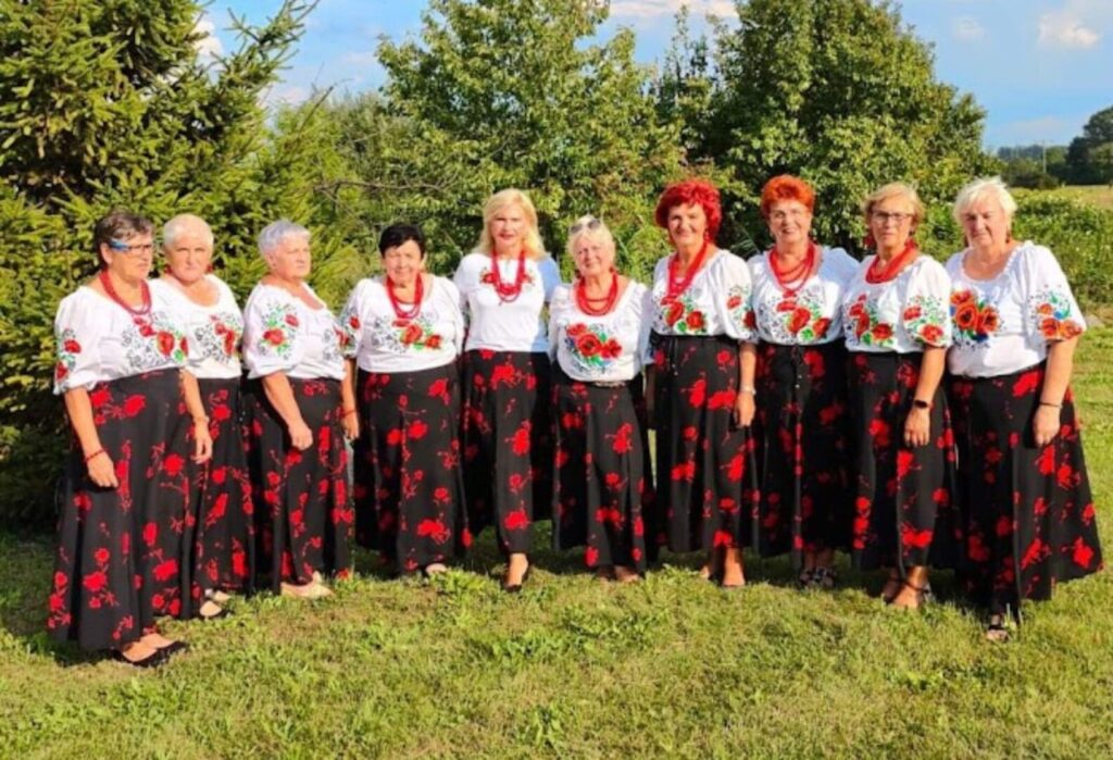Grupa seniorek w ludowych strojach pozuje wspólnie na tle zieleni w słoneczny dzień.