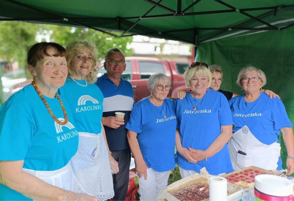Grupa uśmiechniętych seniorów pod zielonym namiotem serwująca domowe ciasta.