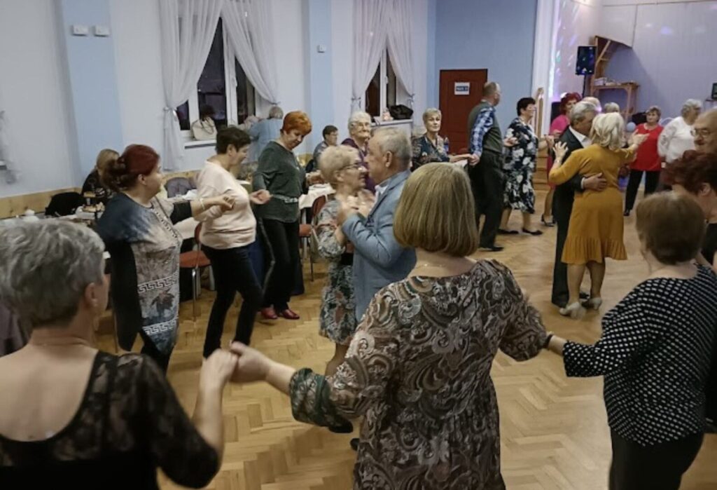 Seniorzy tańczący wspólnie w jasnej sali podczas wieczornej imprezy integracyjnej.