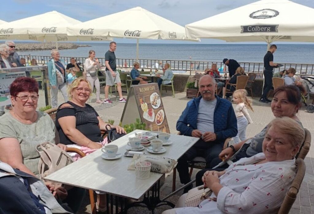 Grupa seniorów siedzi przy stoliku w nadmorskiej kawiarni, w tle widok na morze i deptak.