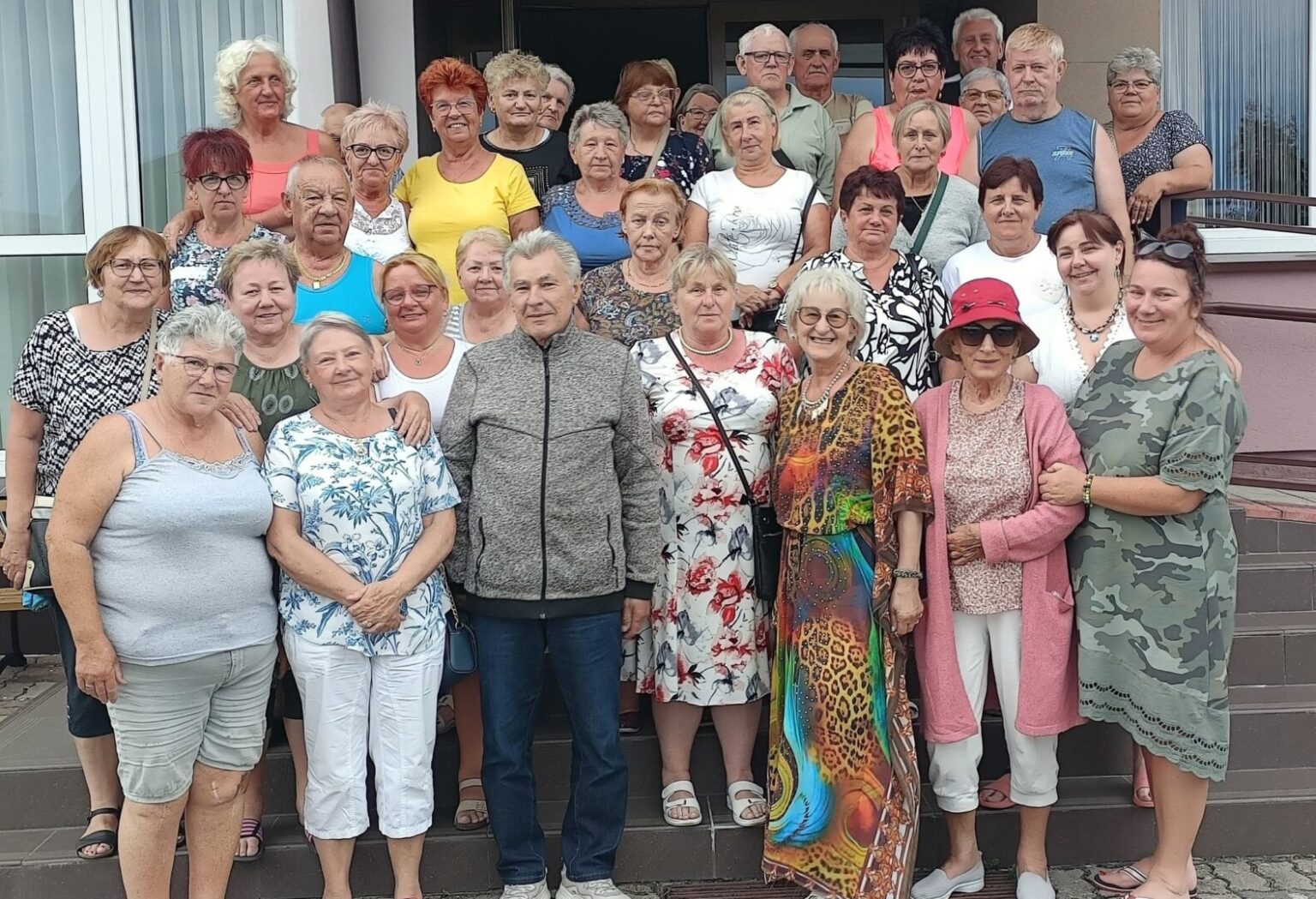Grupa seniorów i seniorek pozuje wspólnie na schodach przed budynkiem wypoczynkowym.
