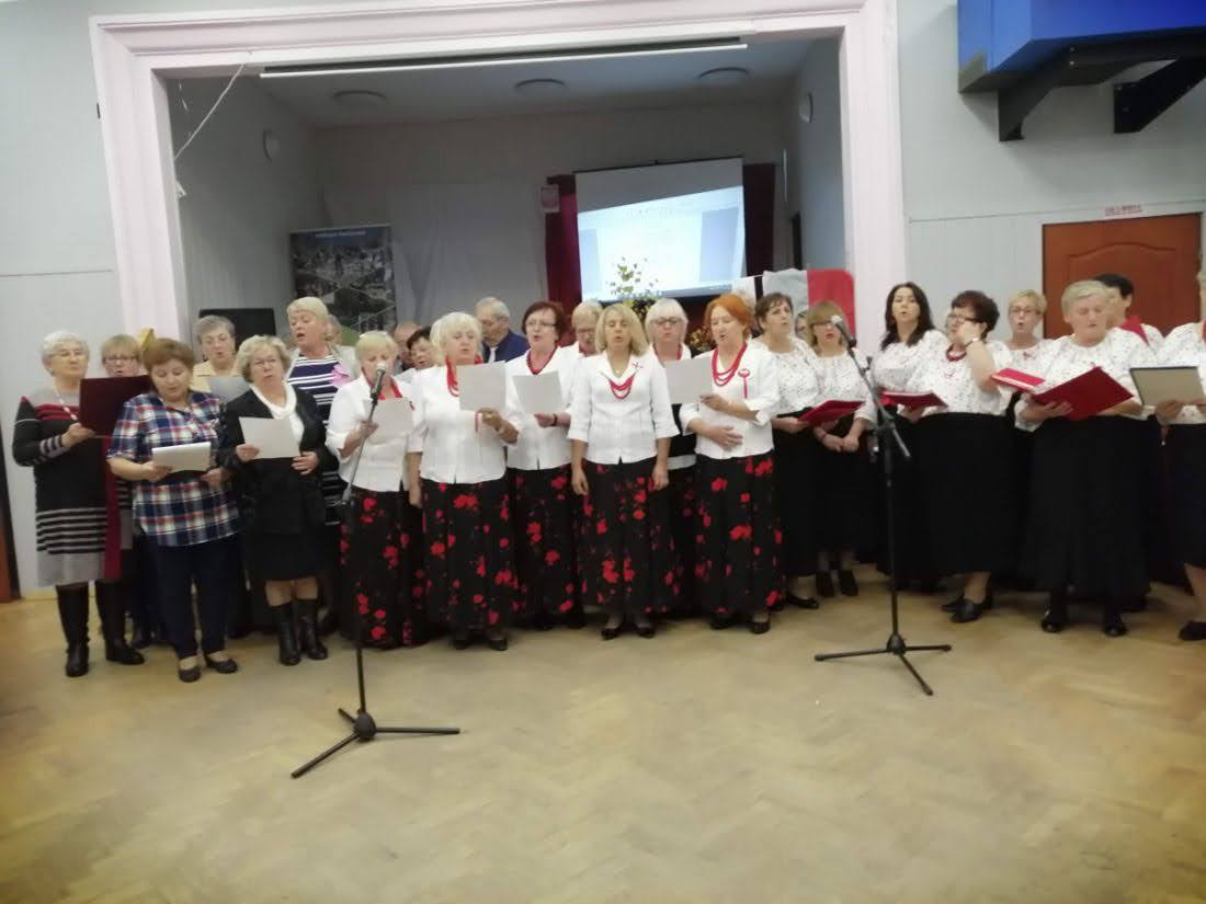 Grupa seniorek i seniorów śpiewa na scenie podczas wydarzenia kulturalnego, stojąc w półokręgu przed mikrofonami.