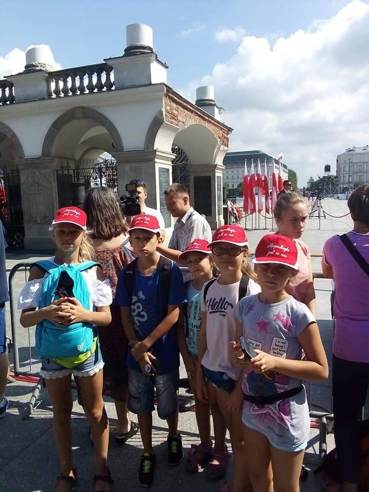 Grupa dzieci w czerwonych czapkach z napisem „Śpiewajmy Patriotycznie” stoi przed Grobem Nieznanego Żołnierza w Warszawie.