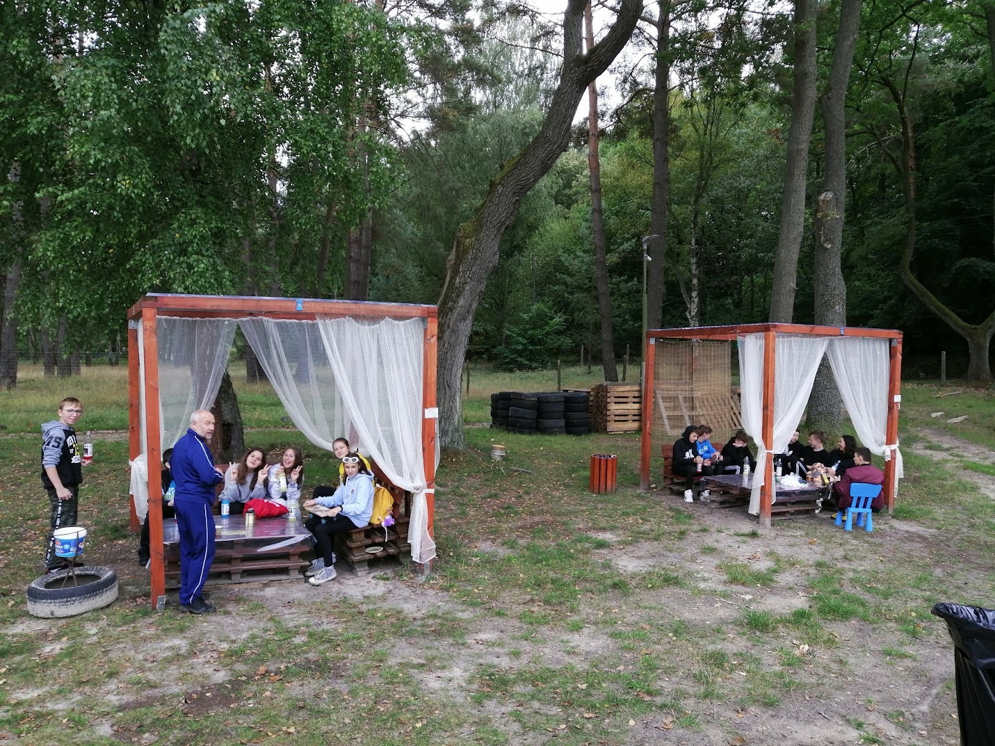 Dwie grupy osób siedzące w altanach z zasłonami, ustawionych na polanie w lesie, biorą udział w plenerowym spotkaniu.