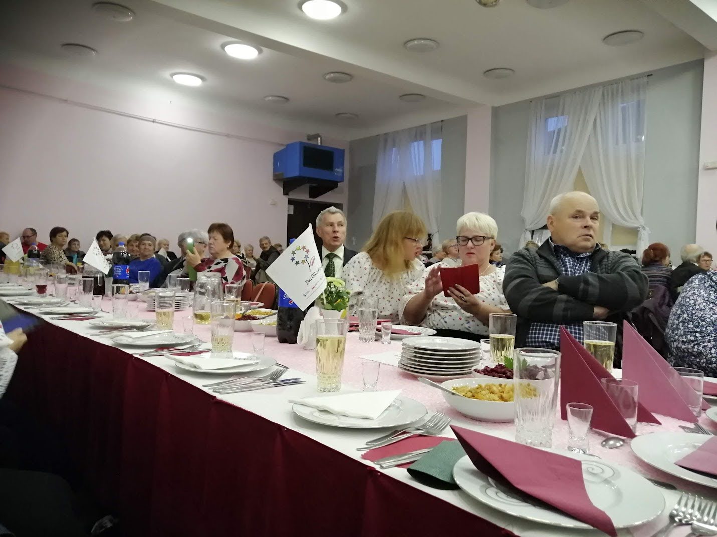 Sala pełna osób starszych uczestniczących w uroczystym spotkaniu przy długim stole nakrytym do wspólnego posiłku.