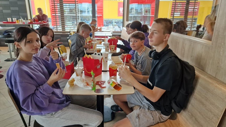 Grupa młodzieży siedzi przy stole w restauracji typu fast food, jedząc wspólny posiłek i rozmawiając.