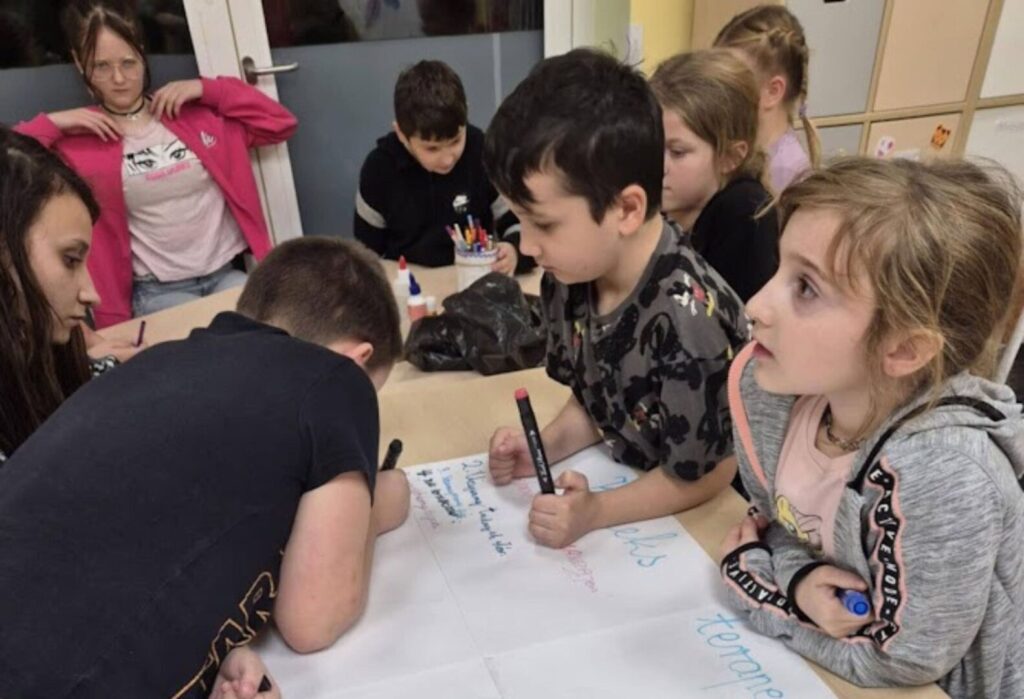 Dzieci pracujące wspólnie przy stole nad dużą kartką papieru, zapisując swoje pomysły markerami podczas zajęć w świetlicy.