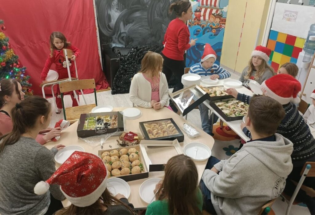 Grupa dzieci i opiekunów w świątecznych czapkach Mikołaja siedzi przy stole pełnym potraw, świętując wspólnie w świetlicy.