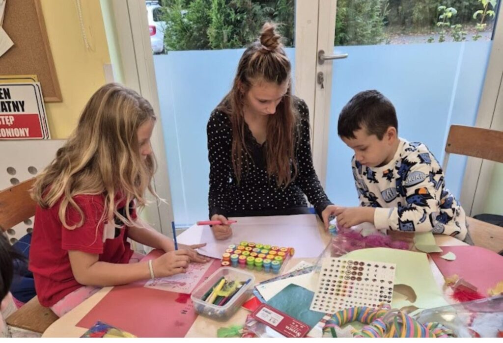 Troje dzieci pracuje nad pracami plastycznymi przy stole, korzystając z farbek, naklejek i kolorowych materiałów w świetlicy.