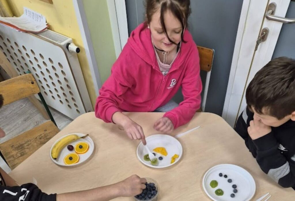 Trójka dzieci układa kompozycje z owoców na papierowych talerzykach podczas zajęć w świetlicy.