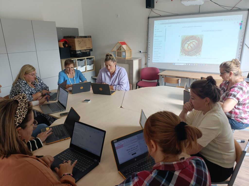 Grupa osób siedzi z laptopami przy stole, w tle wyświetalana jest prezentacja.